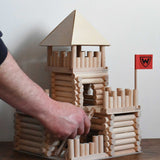 The ladder piece being placed on a fort build created using the Walachia Vario Fort Building Set 194 Pieces. This modular wooden building block system is perfect for those who enjoy a challenge. The sets are available in various sizes at Babipur. 