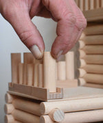 A close up of an adult's hand placing one of the blocks on a base piece to create a fence as part of a fort build created using the Walachia Vario Fort Building Set 194 Pieces. This modular wooden building block system is perfect for those who enjoy a challenge. The sets are available in various sizes at Babipur. 
