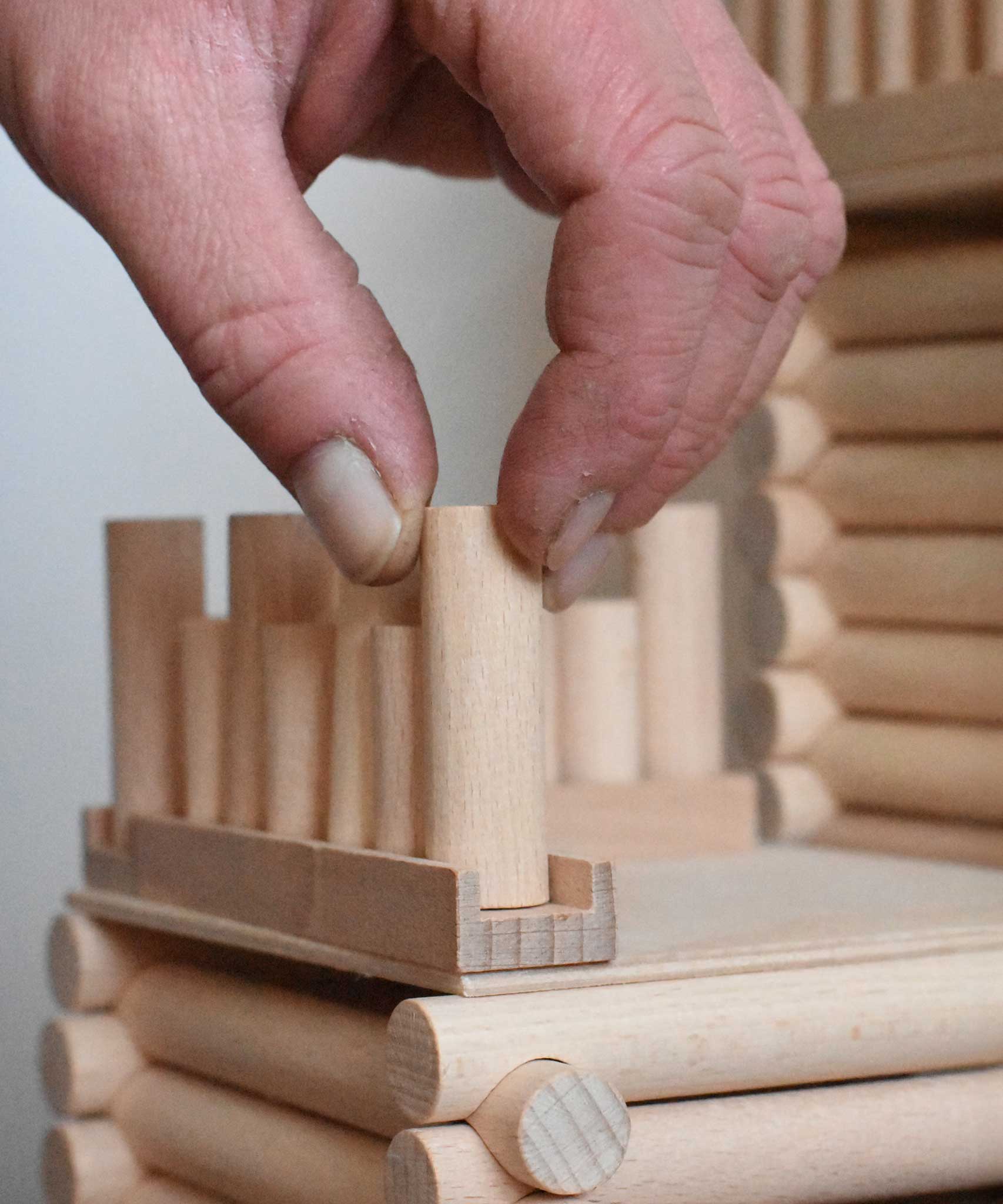 A close up of an adult's hand placing one of the blocks on a base piece to create a fence as part of a fort build created using the Walachia Vario Fort Building Set 194 Pieces. This modular wooden building block system is perfect for those who enjoy a challenge. The sets are available in various sizes at Babipur. 