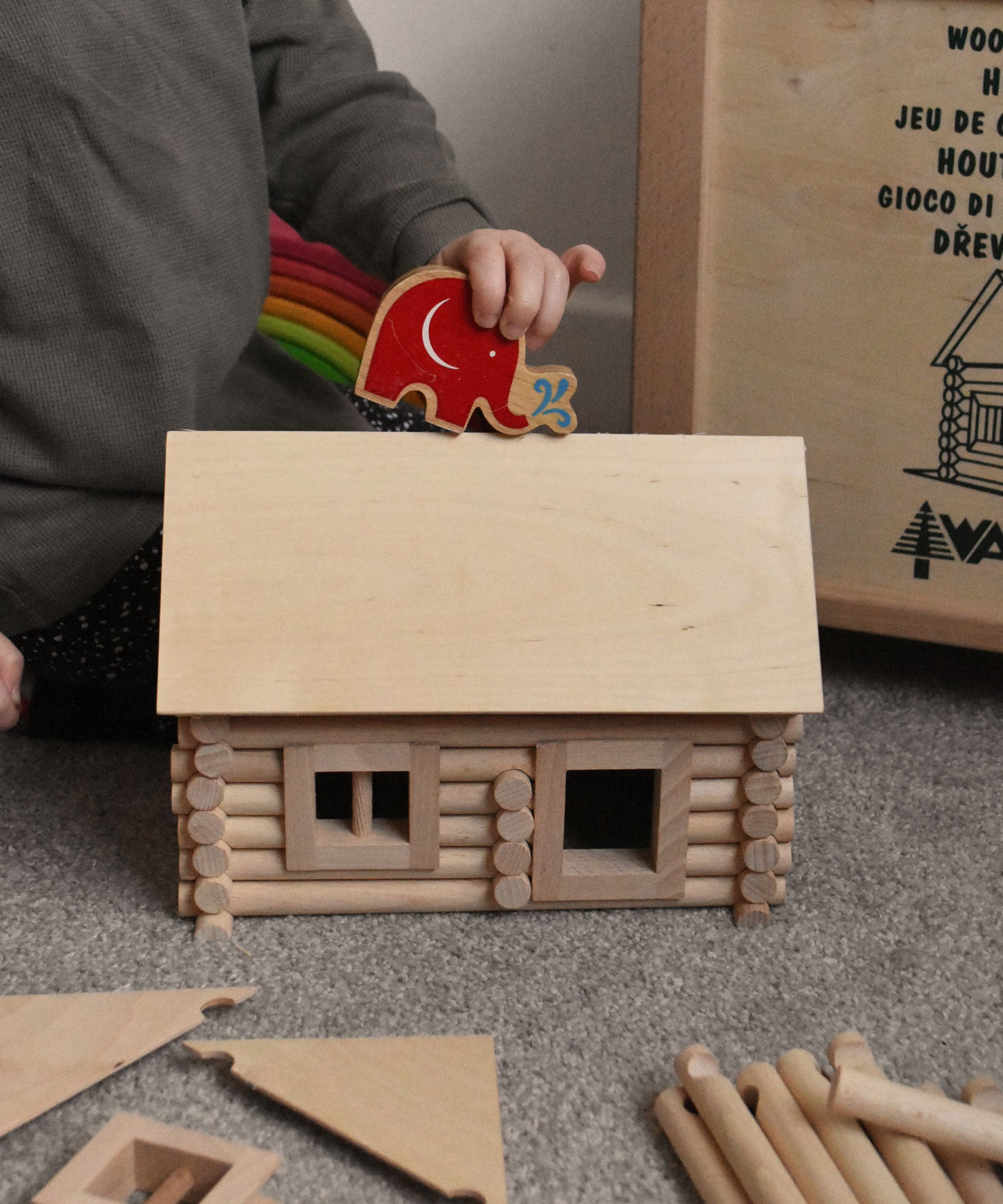 A child placing a Lanka Kade Babipur elephant figure on the roof of a cabin created using wooden blocks from the Walachia Vario Building Sets. This modular wooden building block system is perfect for those who enjoy a challenge. The sets are available in various sizes at Babipur.  