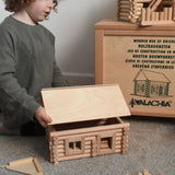 A child placing a roof on a cabin created using wooden blocks from the Walachia Vario Building Sets. This modular wooden building block system is perfect for those who enjoy a challenge. The sets are available in various sizes at Babipur.  