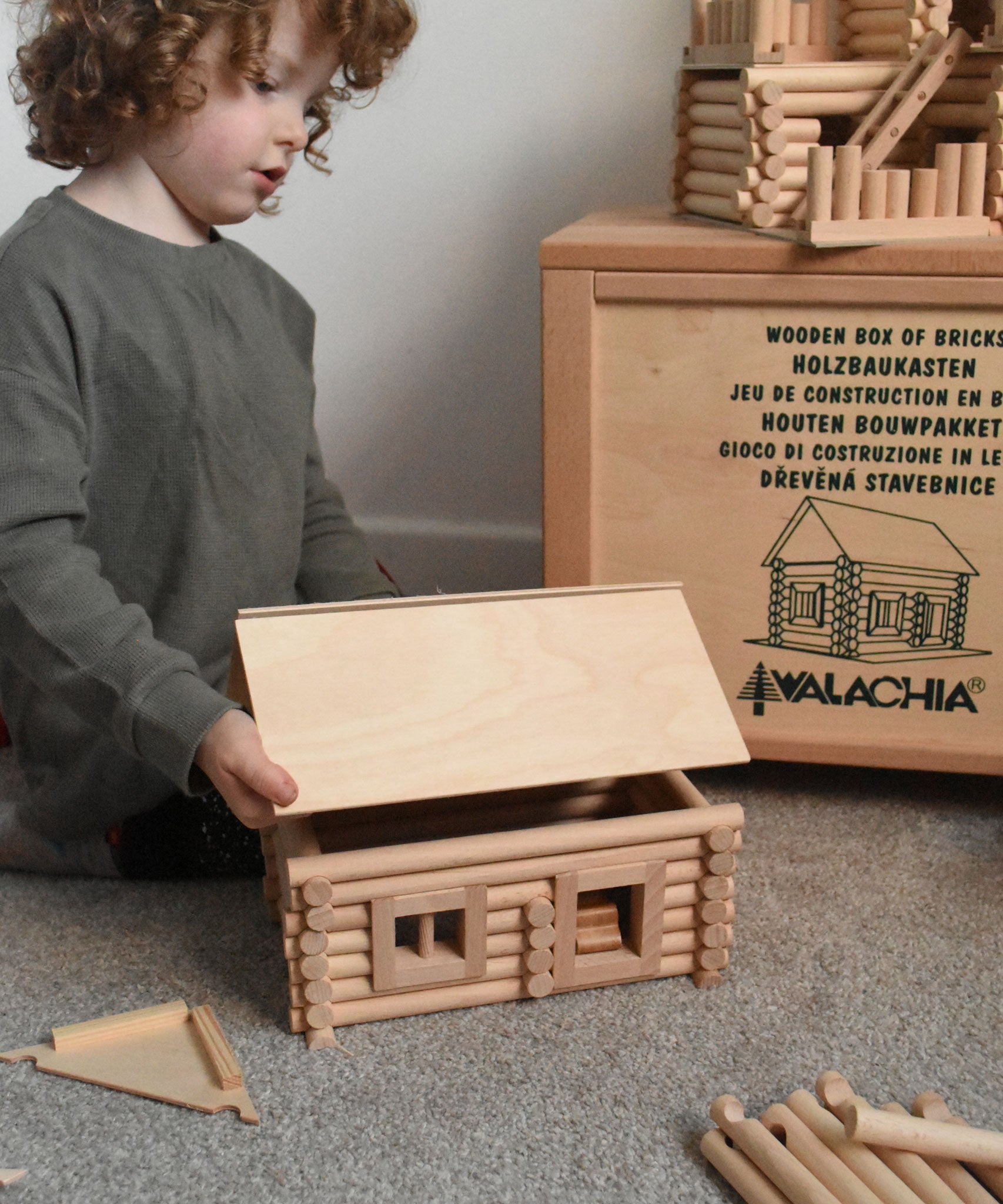 A child placing a roof on a cabin created using wooden blocks from the Walachia Vario Building Sets. This modular wooden building block system is perfect for those who enjoy a challenge. The sets are available in various sizes at Babipur.  