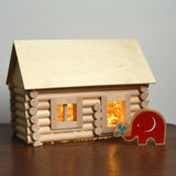 A log cabin created using the Walachia Vario Building Set 72 Pieces.  Fairy lights have been placed inside the cabin and a Lanka Kade Babipur elephant figure is standing outside. This modular wooden building block system is perfect for those who enjoy a challenge. The sets are available in various sizes at Babipur. 