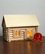 A log cabin created using the Walachia Vario Building Set 72 Pieces.  Fairy lights have been placed inside the cabin and a Lanka Kade Babipur elephant figure is standing outside. This modular wooden building block system is perfect for those who enjoy a challenge. The sets are available in various sizes at Babipur. 