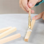 A close up of the gluing process. A child is painting glue onto the wooden sticks to build a WALACHIA wooden house