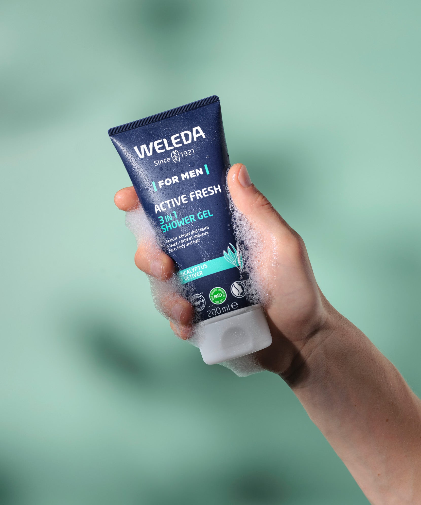 Hand holding a foaming tube of Weleda Men’s Active Fresh 3 in 1 Shower Gel against a green background.