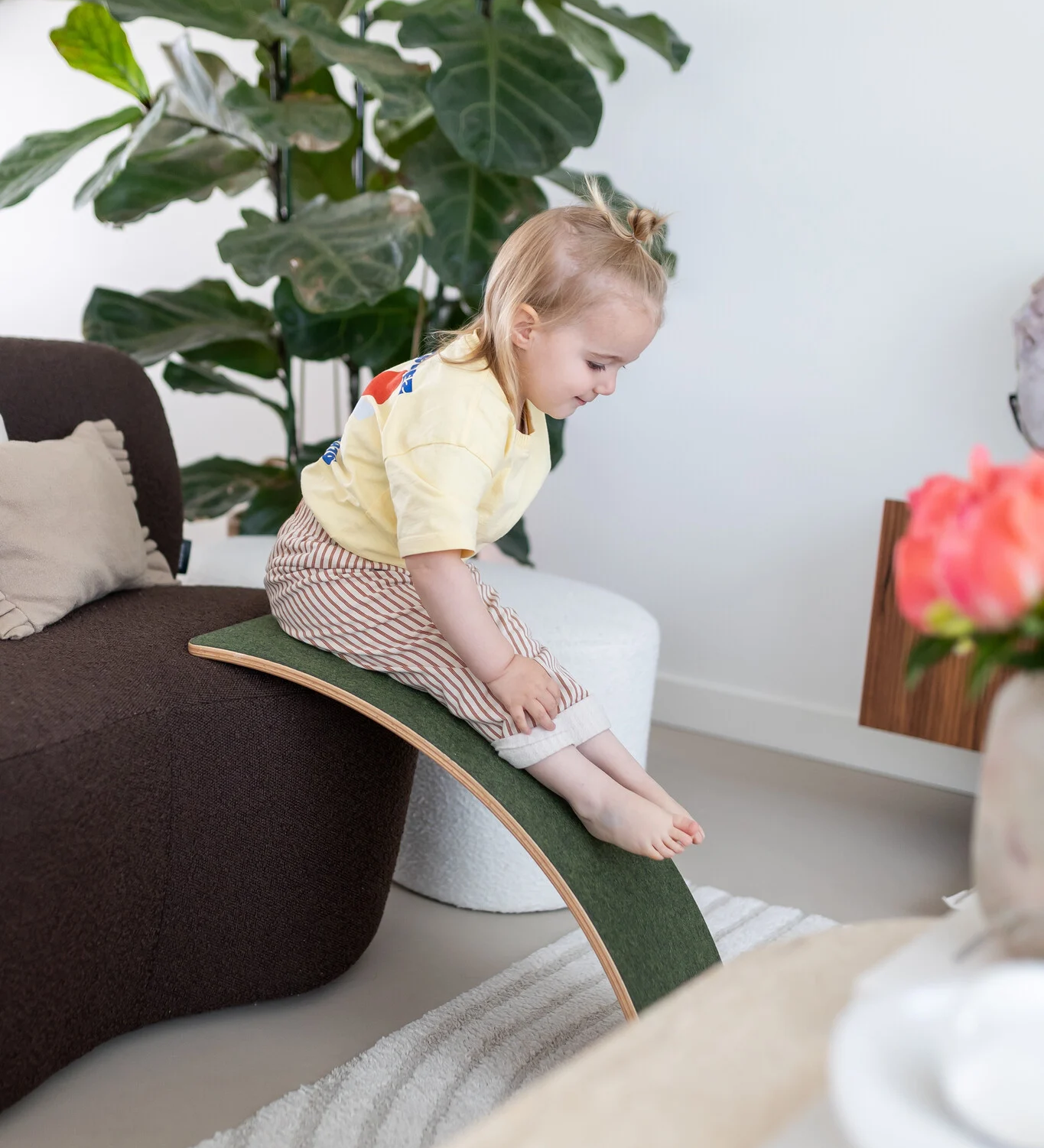 A little toddler sliding down the felt side of a wooden Wobbel Board, leaned against a sofa.