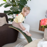 A little toddler sliding down the felt side of a wooden Wobbel Board, leaned against a sofa.