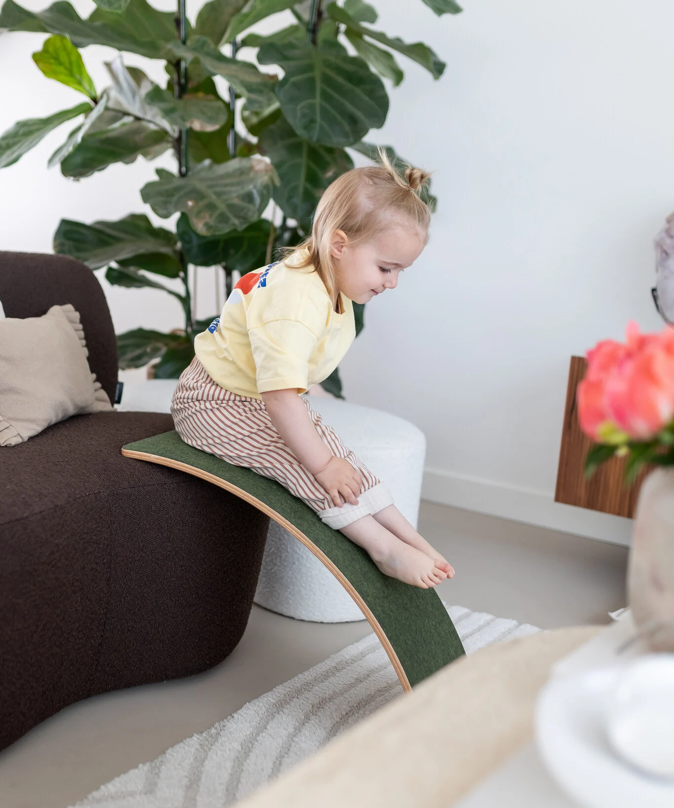 A little toddler sliding down the felt side of a wooden Wobbel Board, leaned against a sofa.