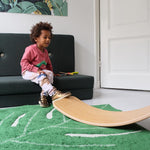 Child resting his foot on a Wobbel Bamboo balance board on a green leaf carpet