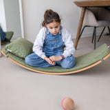 Child sitting on a wooden Wobbel board with a olive green deck on a concrete surface