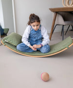 Child sitting on a wooden Wobbel board with a olive green deck on a concrete surface