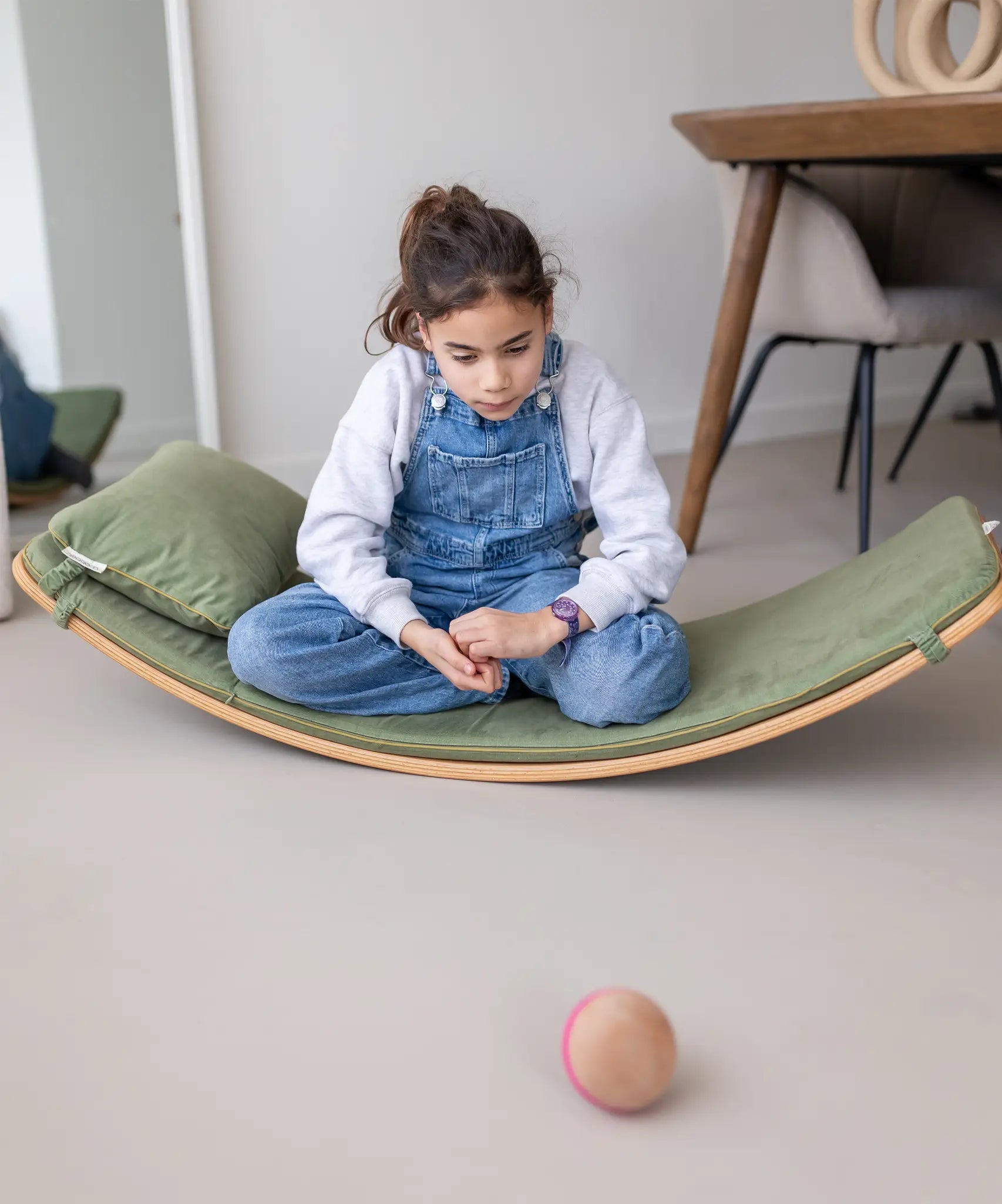Child sitting on a wooden Wobbel board with a olive green deck on a concrete surface