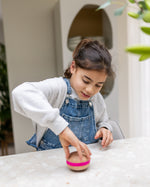 A child playing and spinning a Wooden Wobbel Candy Big Macaron on a  table. Made from Beech wood with a pink "filling"