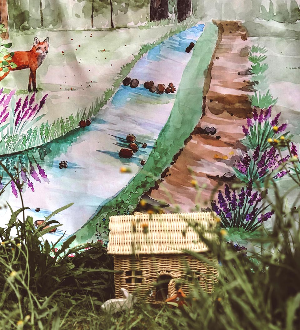 Wondercloths walk in the forest plastic-free play silk hanging up behind a wicker dolls house and some long grass