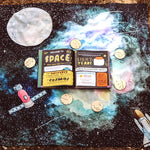 Wondercloths childrens large space print play silk laid out on a woodland floor with a space book on top