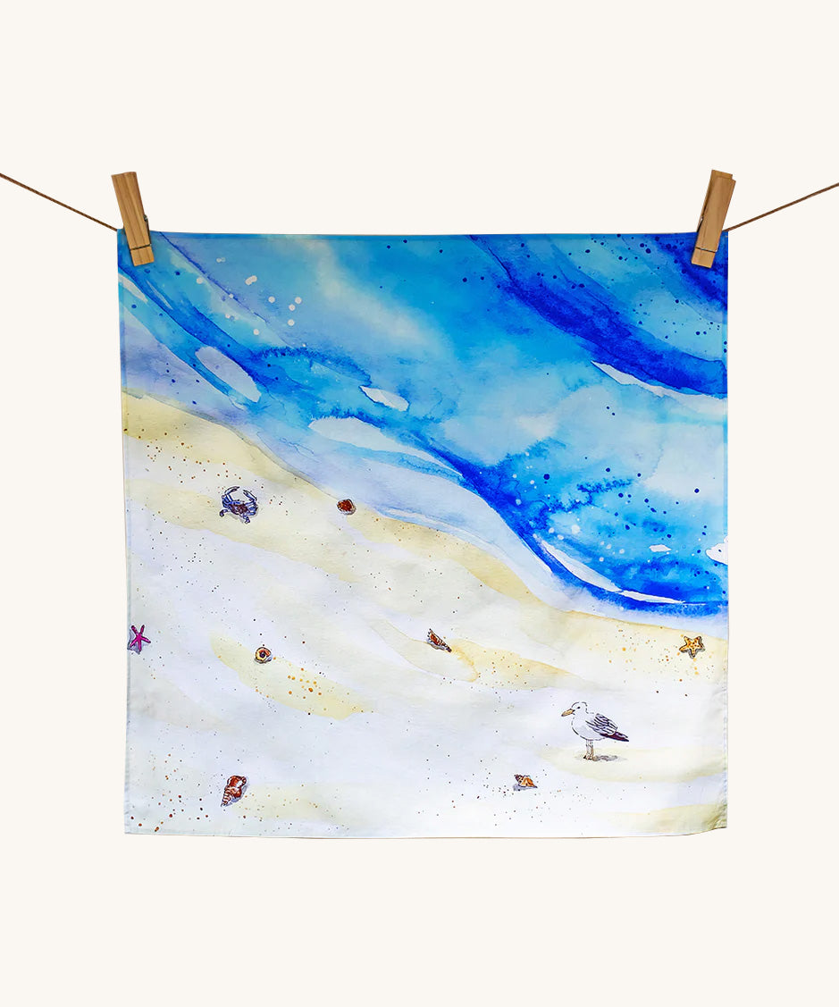 Wonderie a day at the beach playcloth. A watercolour style illustration of the sea and sand with a crab and seagull standing on the and surrounded by shells and starfishes