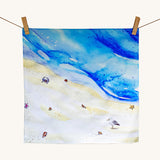 Wonderie a day at the beach playcloth. A watercolour style illustration of the sea and sand with a crab and seagull standing on the and surrounded by shells and starfishes