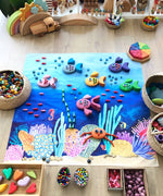 Wonderie Amongst Corals wondercloth play cloth set up on the floor with wooden toys. Fish have been created with Grimm's pebbles and blocks and placed on the cloth.