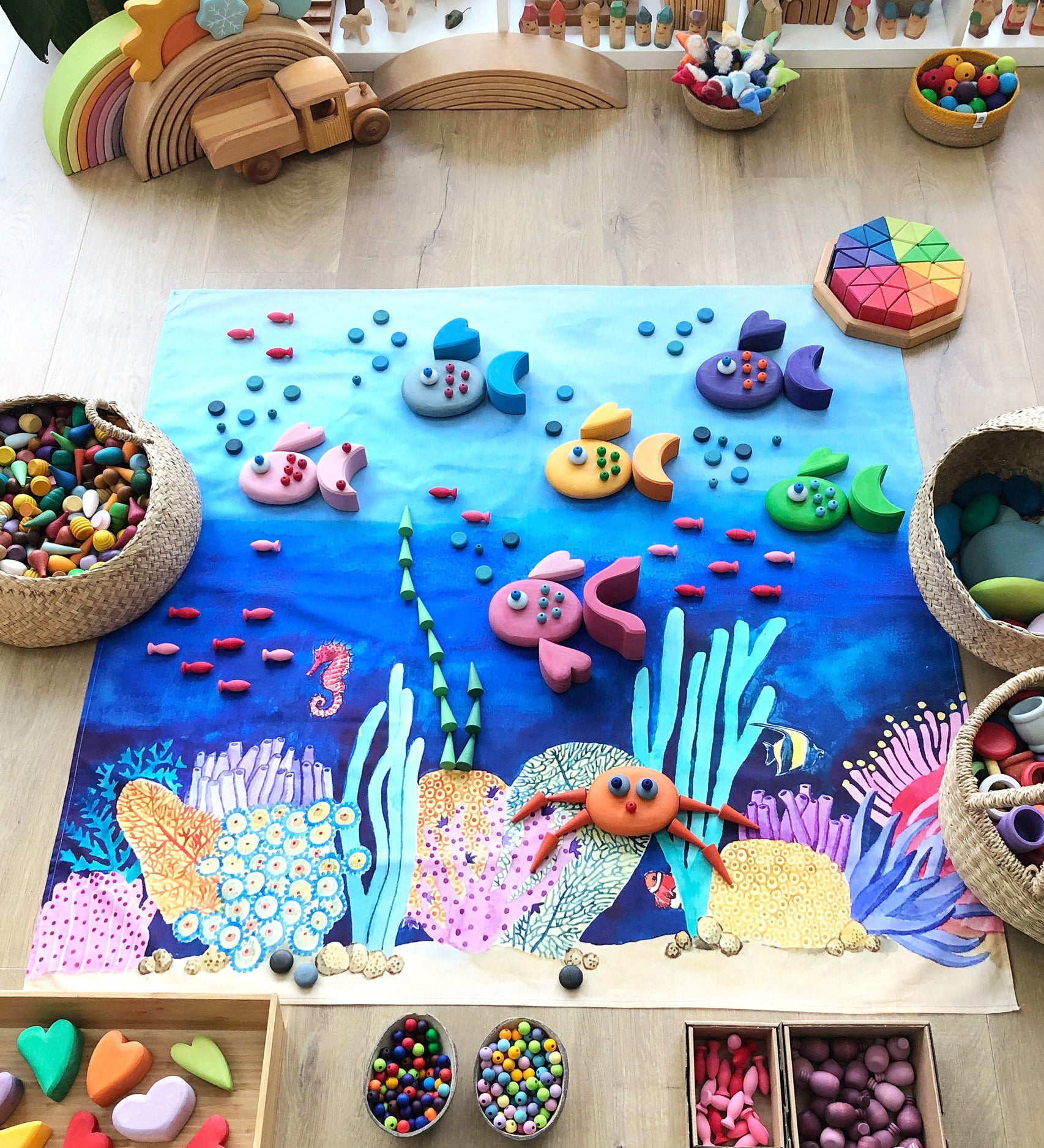 Wonderie Amongst Corals wondercloth play cloth set up on the floor with wooden toys. Fish have been created with Grimm's pebbles and blocks and placed on the cloth.