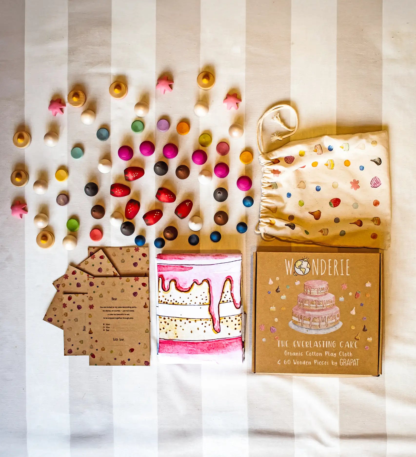 Flatlay showing the contents of the Wonderie play cloth the everlasting cake play set. A square cloth with an iced three tier cake illustration and a set of Grapat loose parts to decorate it with along with a cloth storage bag and party invites