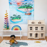 The Wonderie The Lost Island design play cloth hung up on the wall in a playroom. The PlanToys Victorian dolls house, Magic Wood marble tree and the Gwawr rainbow are placed in front. An Olli Ella doll can be seen sitting on the floor at the front on a rug. 