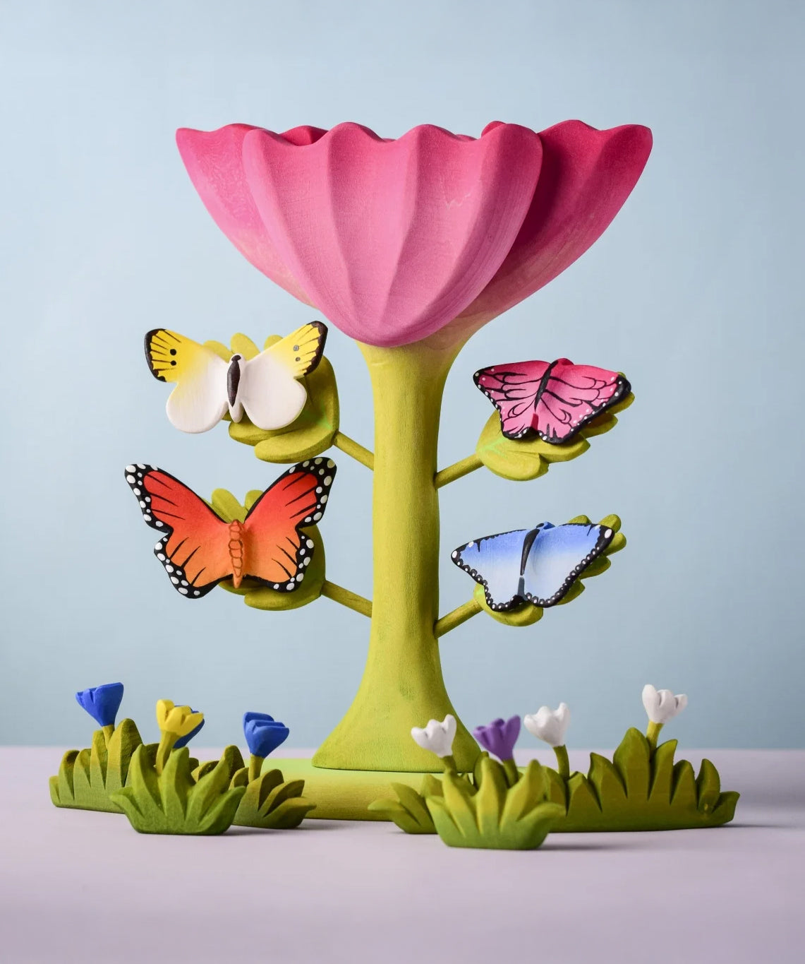Bumbu Toys wooden Butterflies placed on a Bumbu Pink Flower, surrounded by Bumbu flowers and grass.
