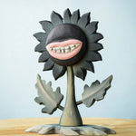 Bumbu Toys black wooden sunflower figure with a mouth with light pink lips and white teeth carved into it for a creepy Halloween vibe. The toy sunflower figure is placed on a wooden surface. 