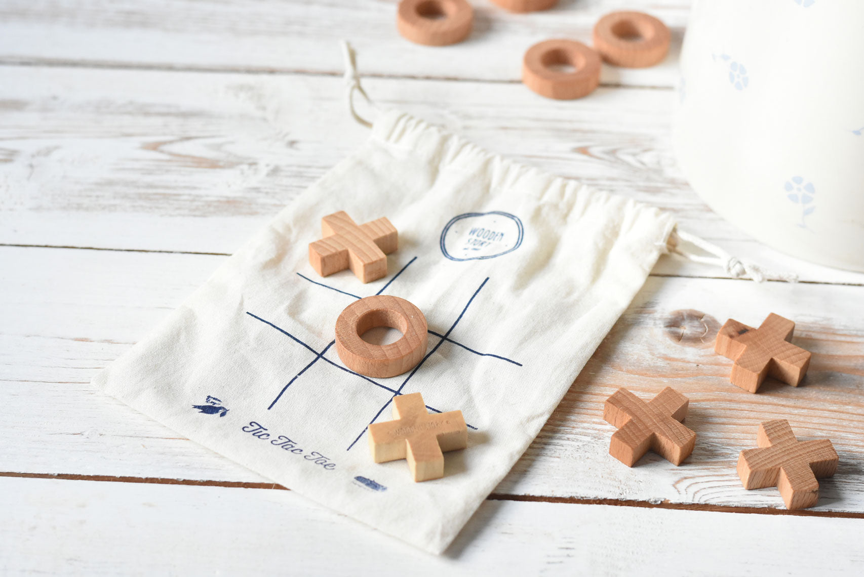The Cotton drawstring storage bag placed on a white wooden surface with pieces from the Wooden Story Tic Tac Toe game placed on the printed grid on the bag. 
