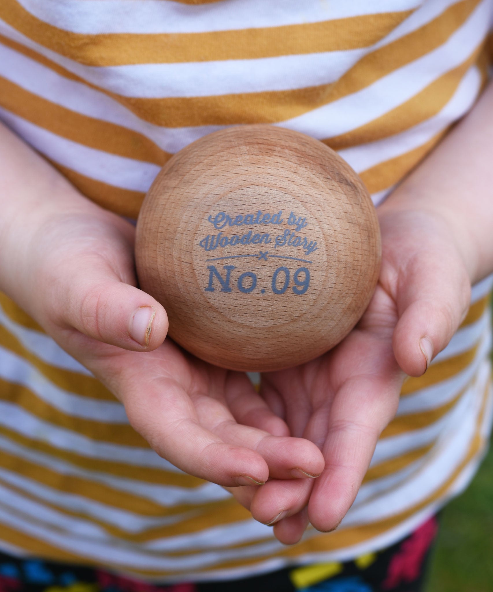 A close of of a wooden boules ball in a child's hand. The words 'created by Wooden Story and No.9 can be seen printed in grey on the ball. 