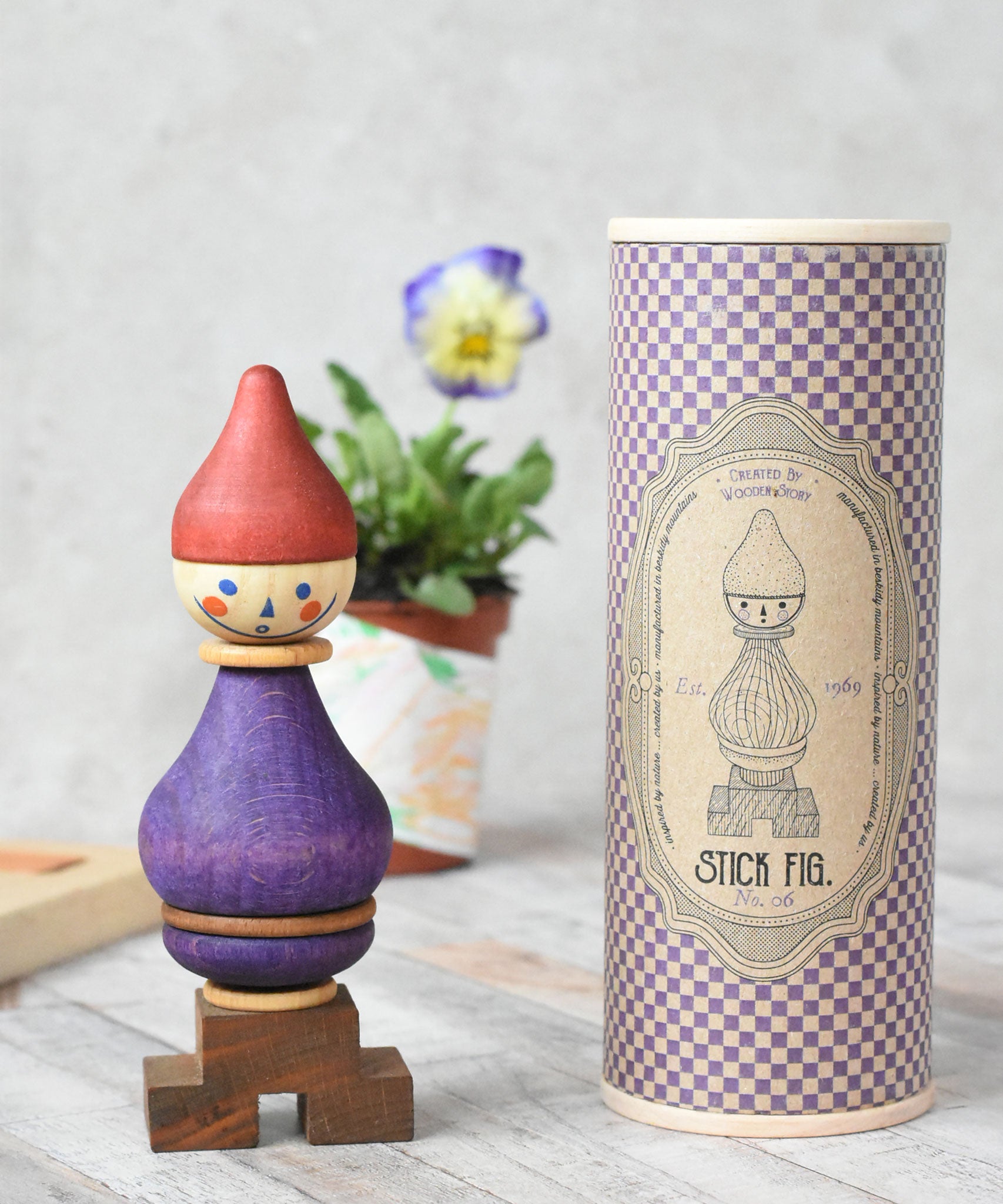 The Wooden Story Stacking Toy Stick Purple Number 6 Figure pictured next to the beautifully patterned tube shaped packaging.  