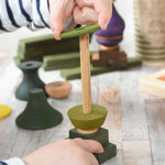 A close up of a child playing with the Wooden Story green stick figure stacking toy, the child is lifting pieces off the stack's base piece. There is a base which is the figures legs and a fixed wooden pole which the other pieces stack onto.