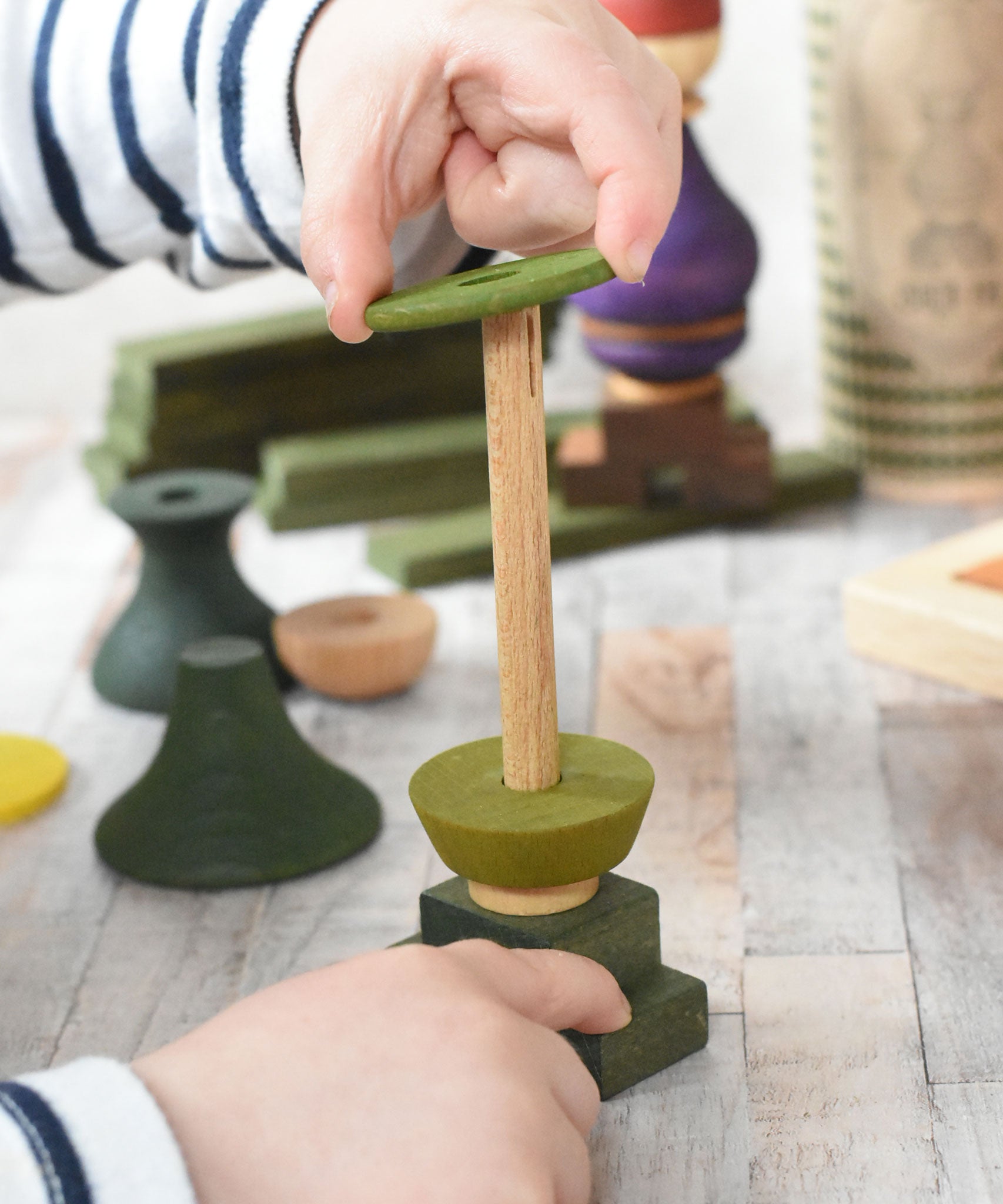 A close up of a child playing with the Wooden Story green stick figure stacking toy, the child is lifting pieces off the stack's base piece. There is a base which is the figures legs and a fixed wooden pole which the other pieces stack onto.