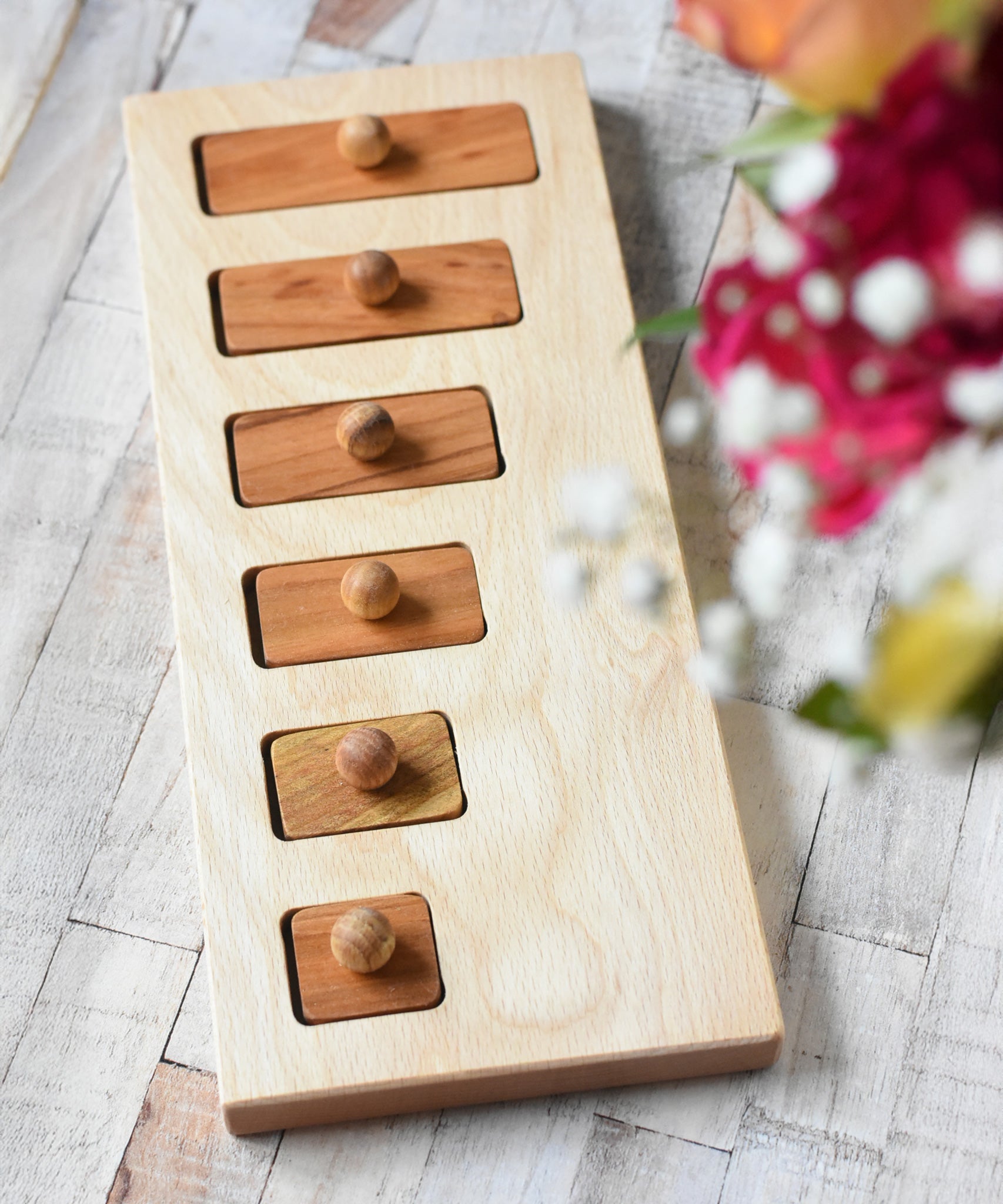 The Wooden Story long short traditional style puzzle toy placed on a wooden surface, flowers can be seen in the foreground. 