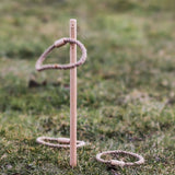 The Wooden Story Loop Toss game set up on grass outdoors. One loop can be seen mid air with two other laying on the grass besides the stick.