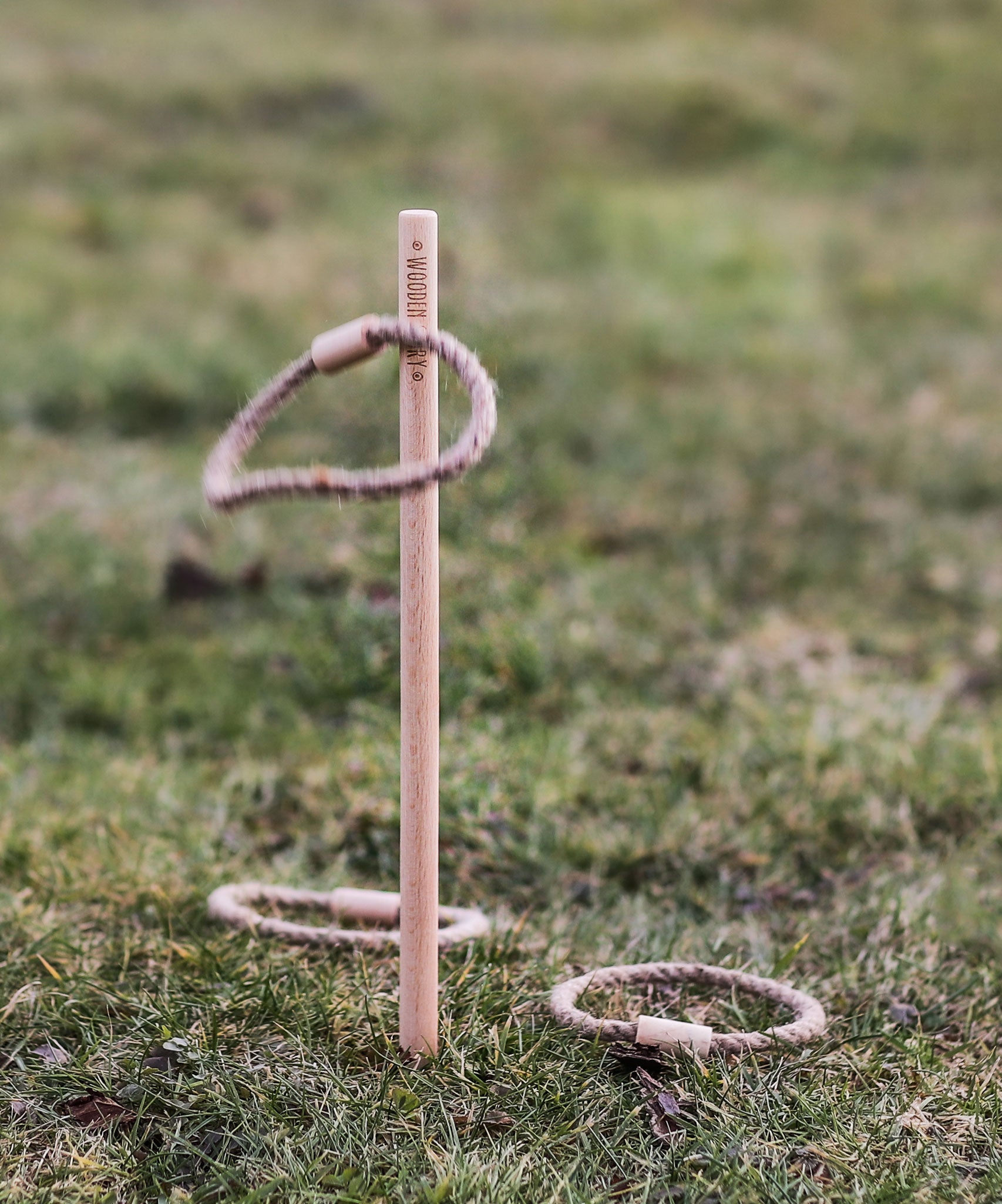The Wooden Story Loop Toss game set up on grass outdoors. One loop can be seen mid air with two other laying on the grass besides the stick.