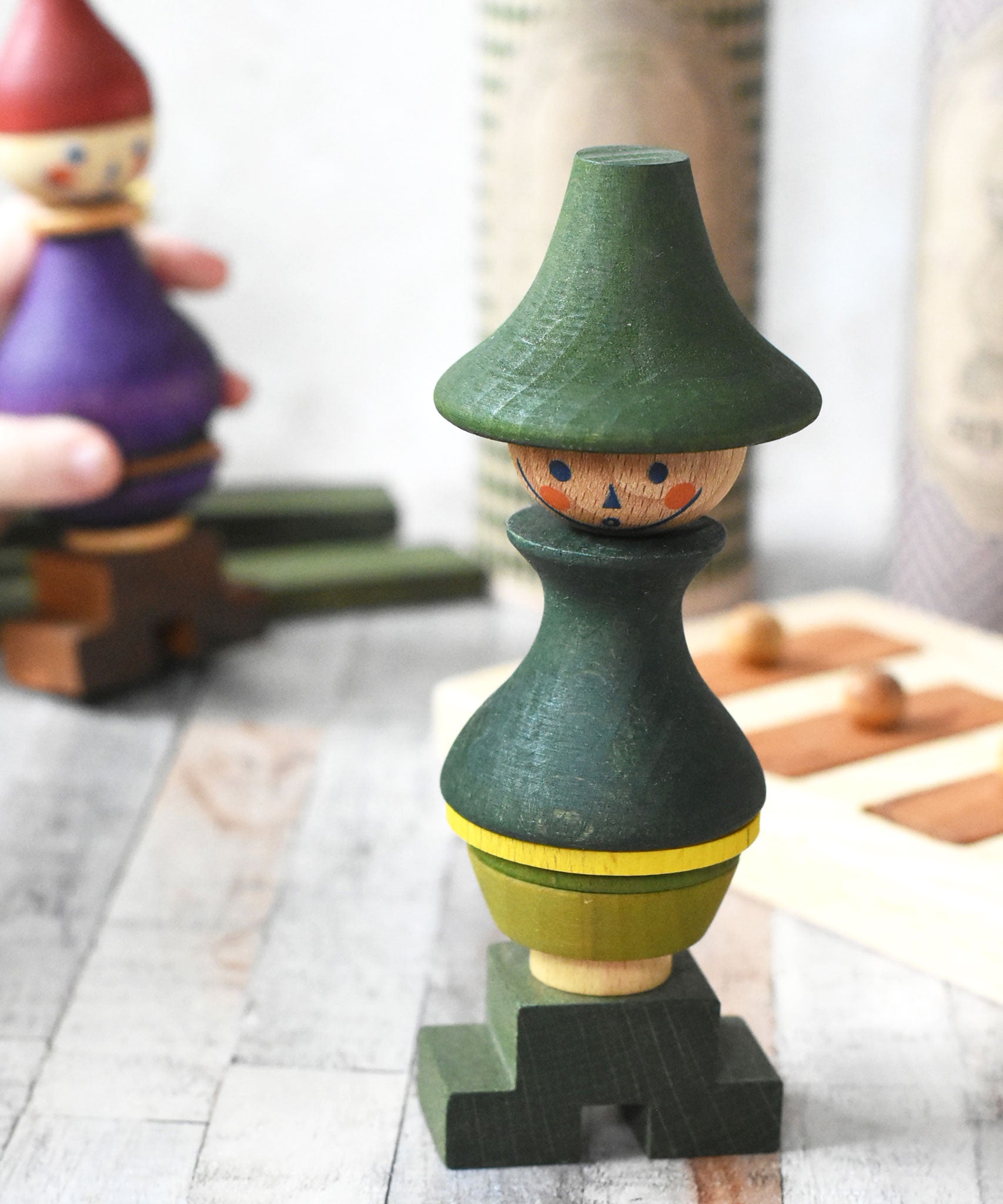 The Wooden Story green stick figure stacking toy on a wooden surface. It's box can be seen in the background along with the purple coloured stick figure. 