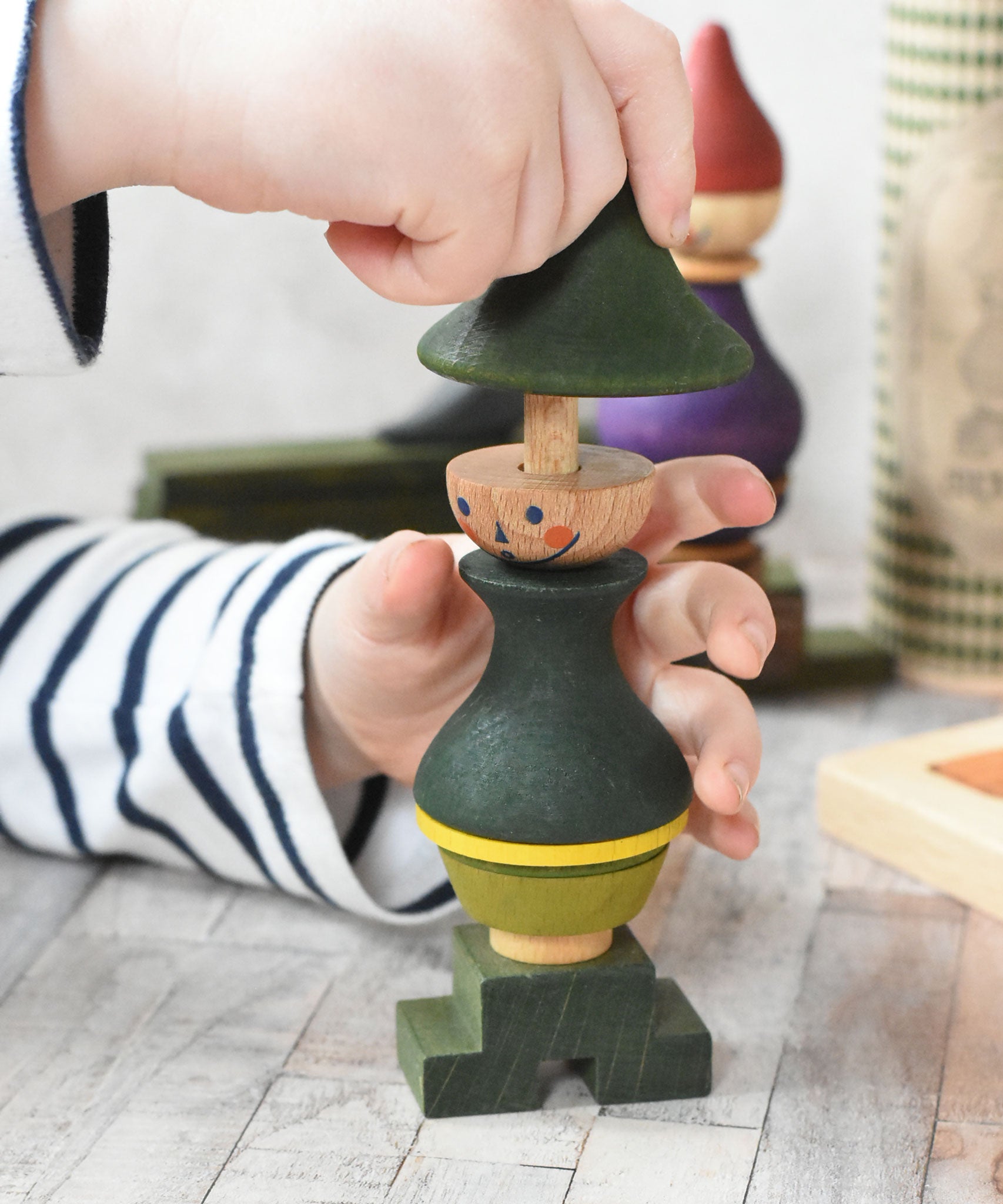 A close up of a child playing with the Wooden Story green stick figure stacking toy, the child is lifting the top, hat shaped piece off the stack.
