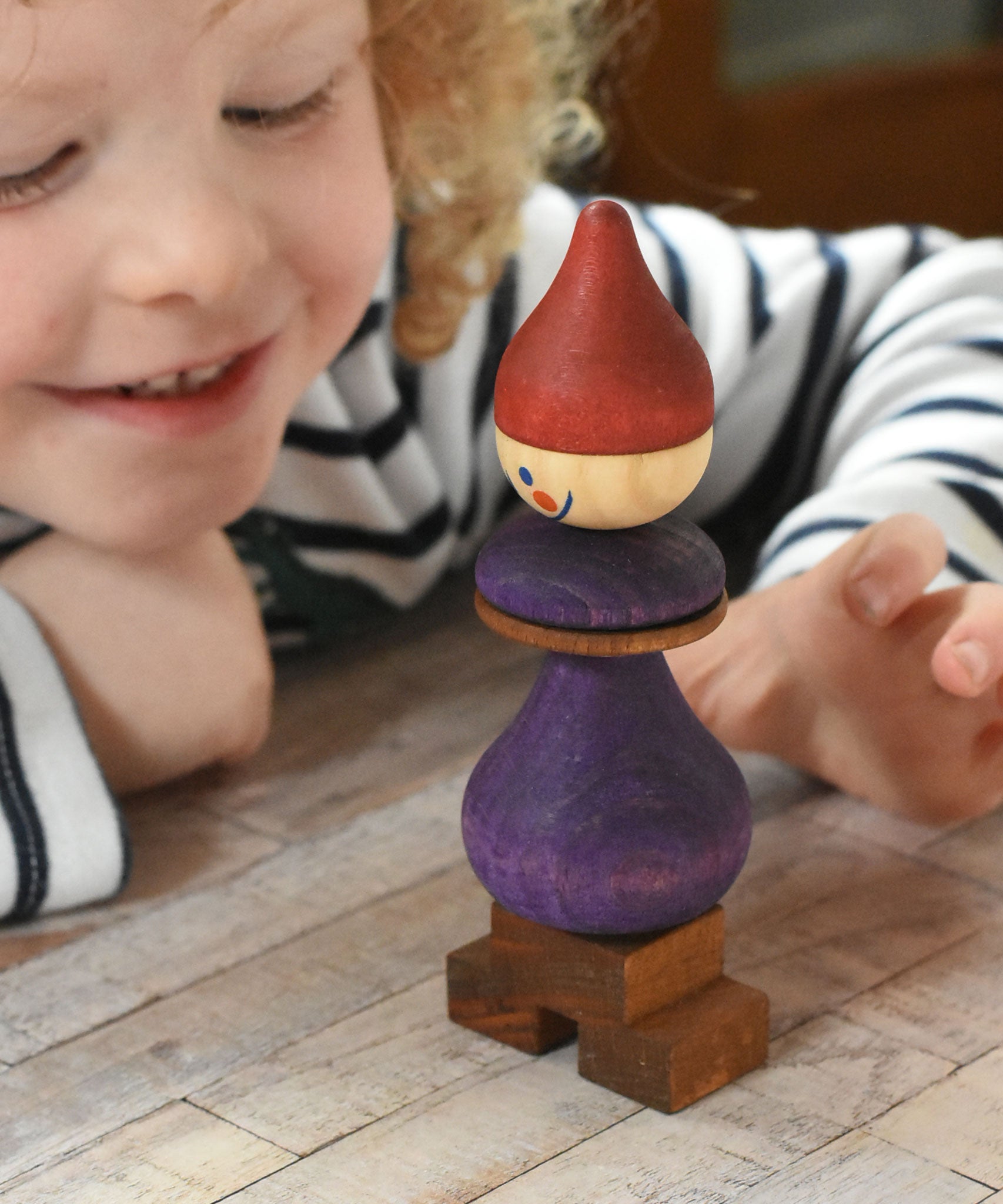 The Wooden Story Stacking Toy Stick Purple Number 6 Figure on a wooden surface. A smiling child can be seen in the background. 