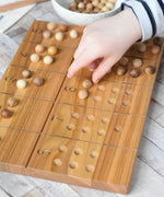 A close up of a child's hand placing wooden balls on the Wooden Story Montessori Number Tracing and Counting Board. The counting side of the board features the numbers from 1 to 10 alongside a corresponding number of indents that the small wooden balls can sit on.