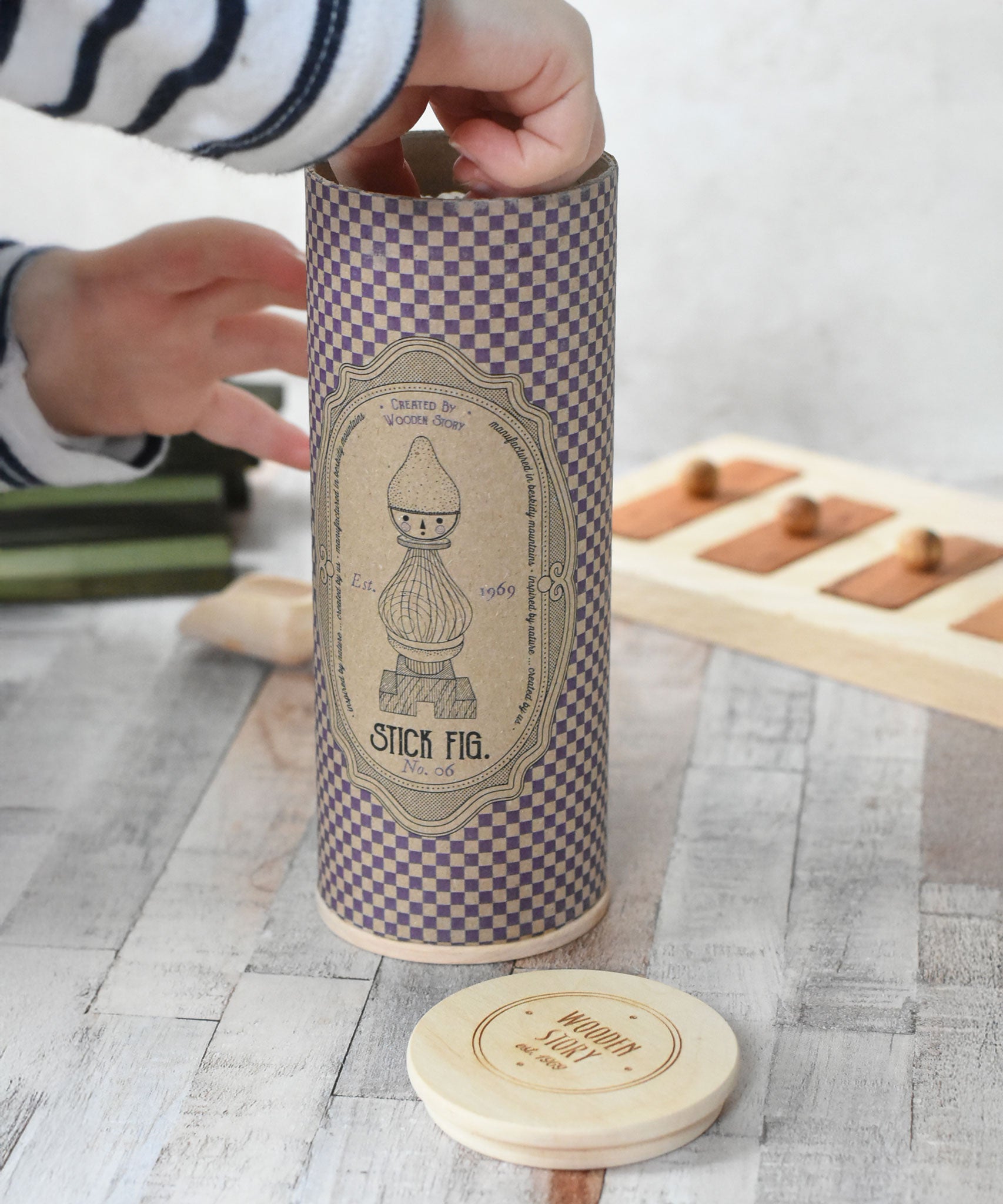 The decorated tube gift box that contains the decorated Wooden Story Stacking Toy Stick Purple Number 6 Figure. The rigid tube has a wooden disc lid.