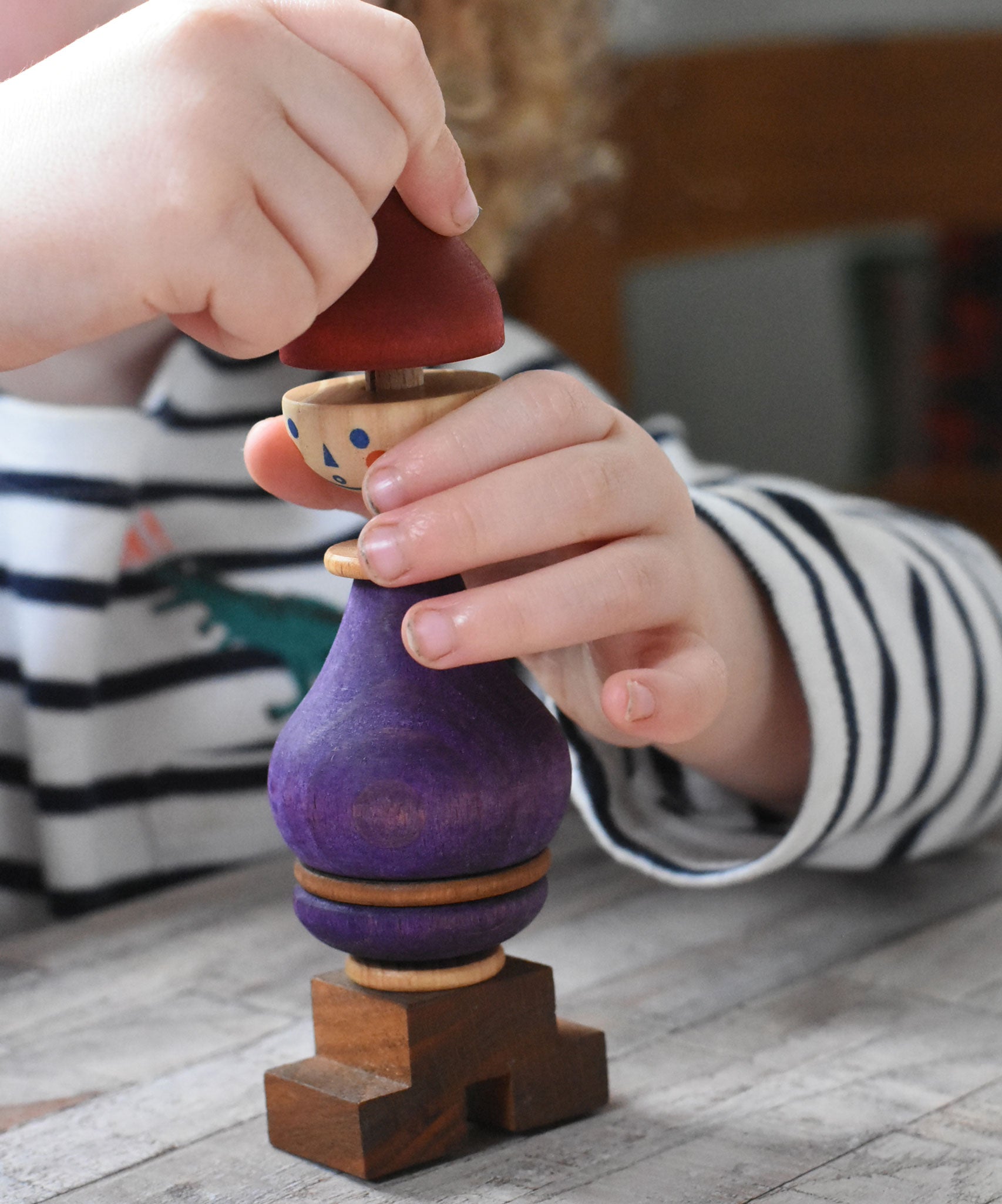 A close up of a child's hand playing with the Wooden Story Stacking Toy Stick Purple Number 6 Figure. They are lifting the maroon red coloured hat shaped piece off the top.