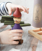 A close up of a child's hand playing with the Wooden Story Stacking Toy Stick Purple Number 6 Figure. They are lifting the maroon red coloured hat shaped piece off the top.
