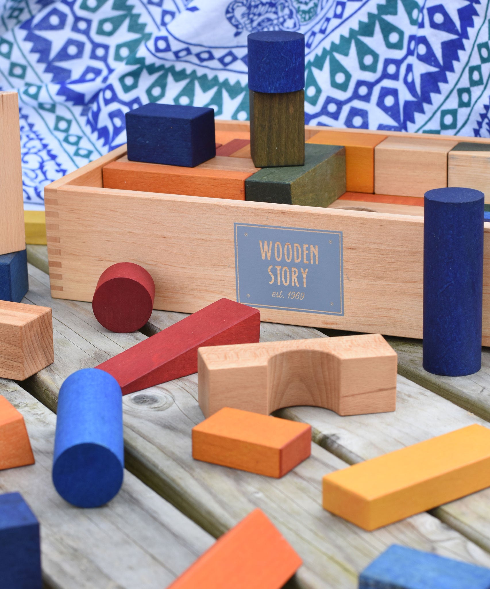 The blocks from the Wooden Story Rainbow XL Blocks set laid out on a wooden surface, the wooden storage box can be seen in the background.