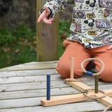 A close up of  the Wooden Story Ring Toss Game shown in play. A child can be seen in the background throwing one of the rings. The ring can be seen mid-air about to land on one of the posts.