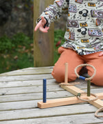 A close up of  the Wooden Story Ring Toss Game shown in play. A child can be seen in the background throwing one of the rings. The ring can be seen mid-air about to land on one of the posts.