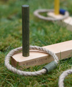 A close up of the Wooden Story logo that's engraved onto one of the base pieces of the  Ring Toss Game. The green stick piece can be seen with a green rope ring on it.