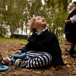A child playing with two blue pieces from the Wooden Story Rainbow Stacker. The child is sitting on a muddy floor outdoors surrounded by fallen leaves and trees. 