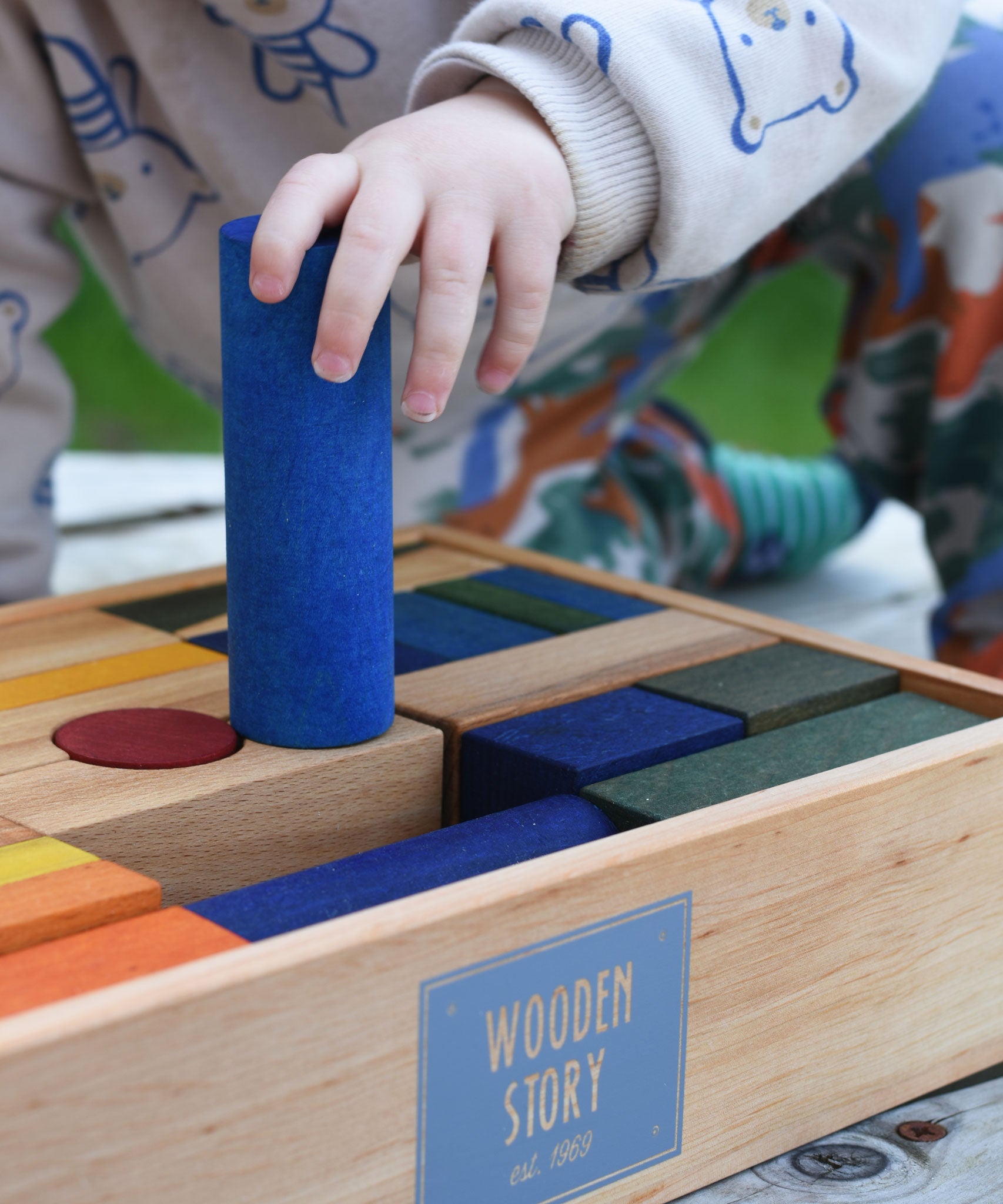 A close up of a child holding the large blue cylinder shaped block from the Wooden Story Rainbow XL Blocks set in their hand. 