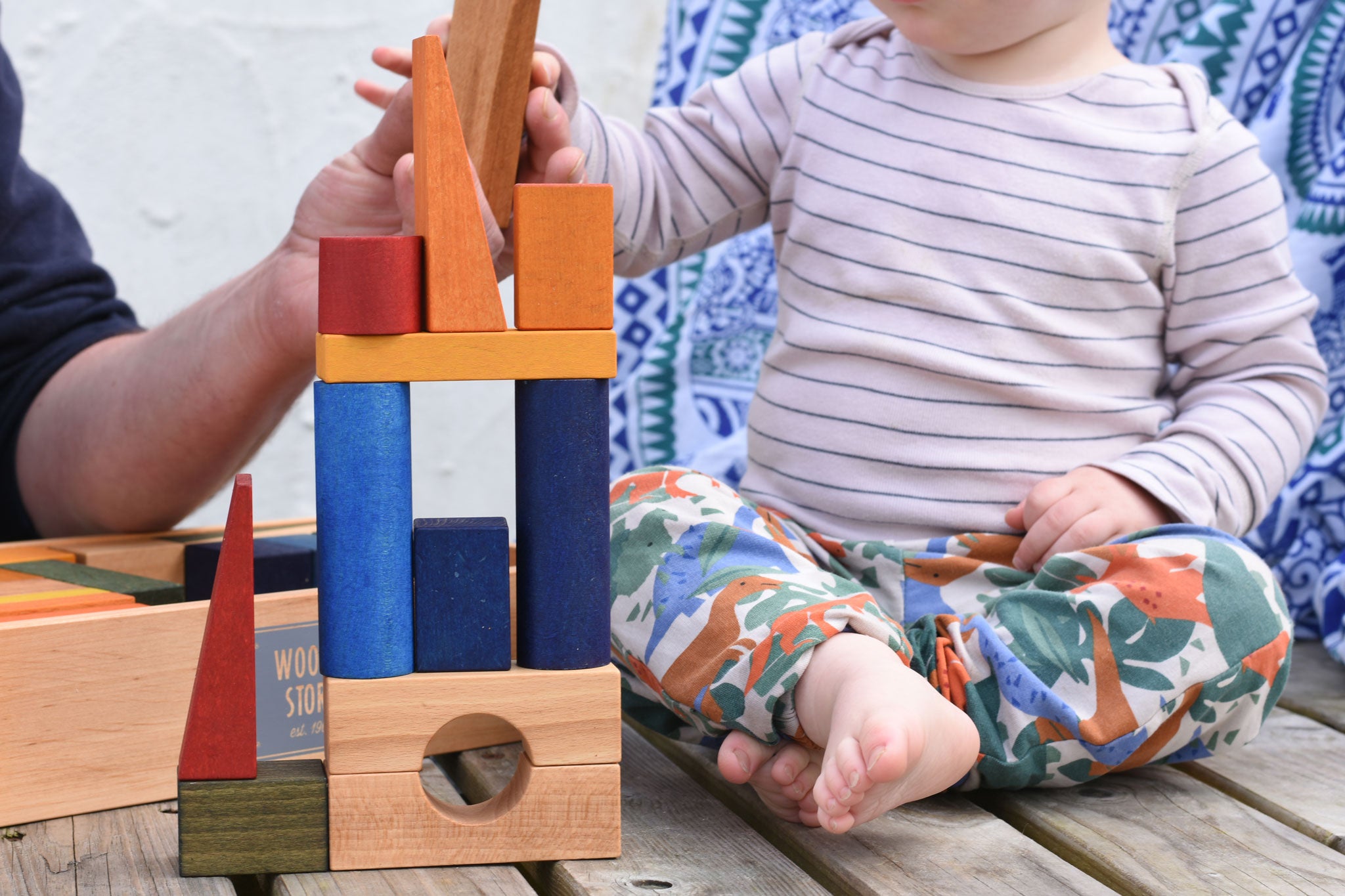 A stack created with the blocks from the Wooden Story Rainbow XL Blocks An adult can be seen in the background passing a natural coloured block over to a young child.